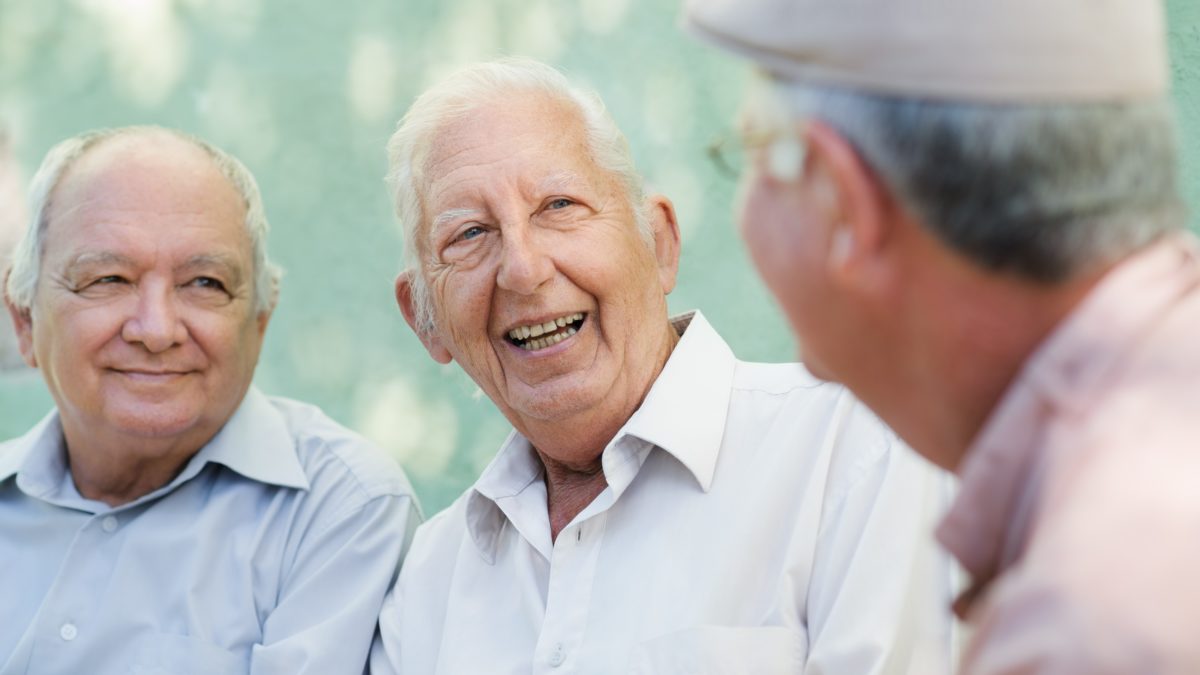 Active retirement, group of three old male friends talking and laughing on bench in public park